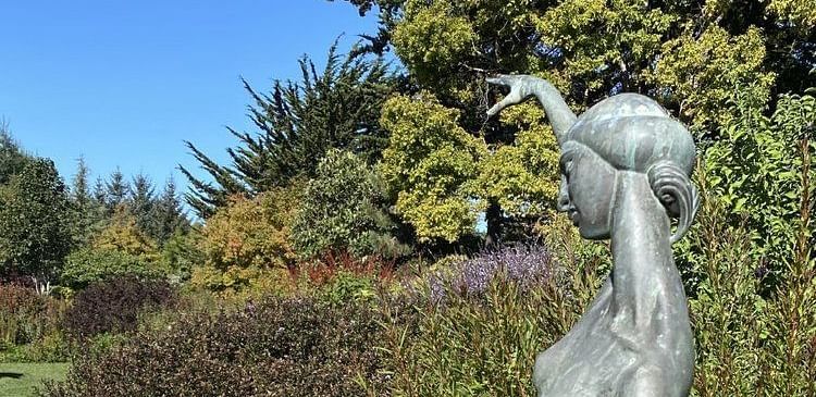 A statue of a heron or similar bird standing on a rock in a garden setting with trees and flowers in the background.