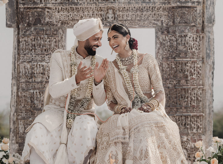 A bride and groom in ivory attire and floral garlands laugh together under a carved stone arch