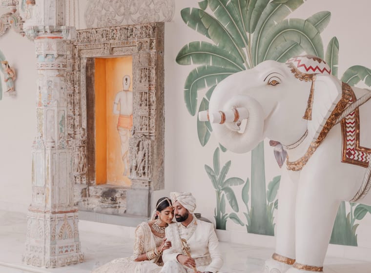 A couple sits beside a white ceremonial elephant statue and a carved stone shrine doorway with painted palm leaves
