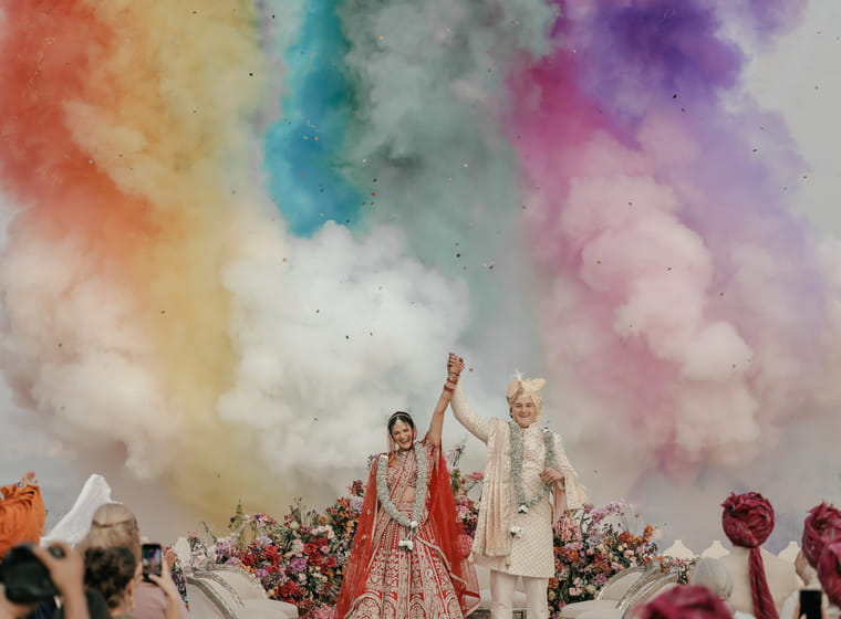 Newlyweds raise their joined hands amid bursts of rainbow-coloured smoke and floral celebration