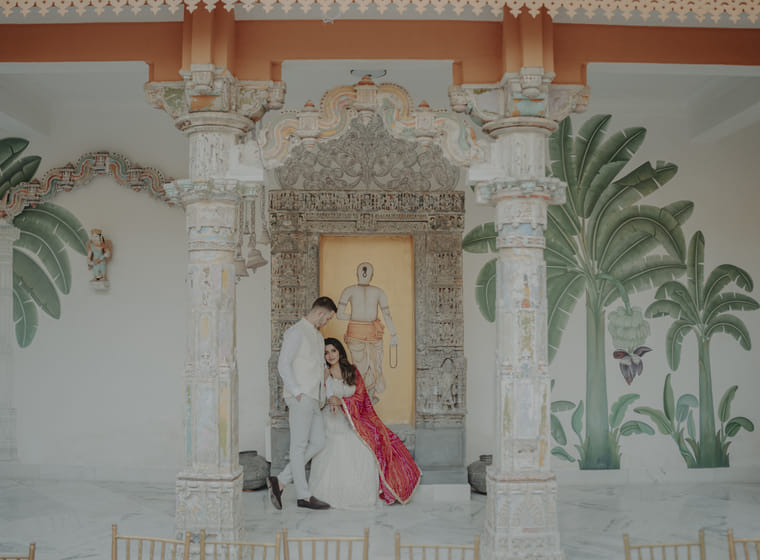 A couple poses beneath an ornately carved heritage archway with hand-painted murals, the bride in white with a red dupatta