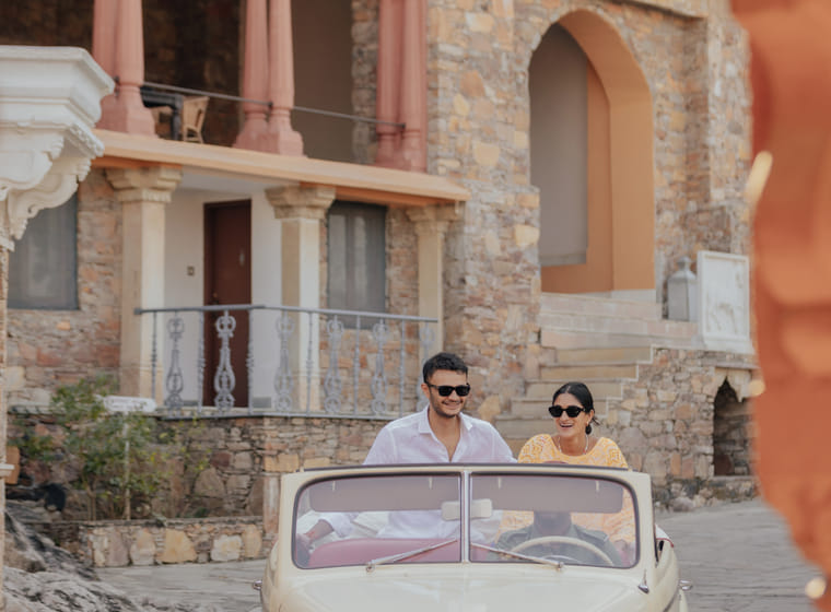 A smiling couple rides in a vintage open-top car outside a heritage stone palace facade