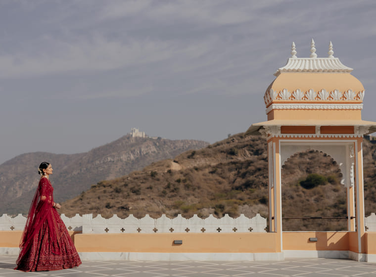 A bride in a red lehenga walks along a rooftop terrace beside a domed pavilion, with hills and a hilltop temple beyond