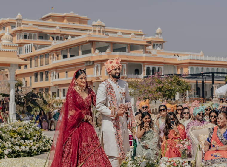 A bride and groom walk down the aisle past seated guests in front of the grand palace facade