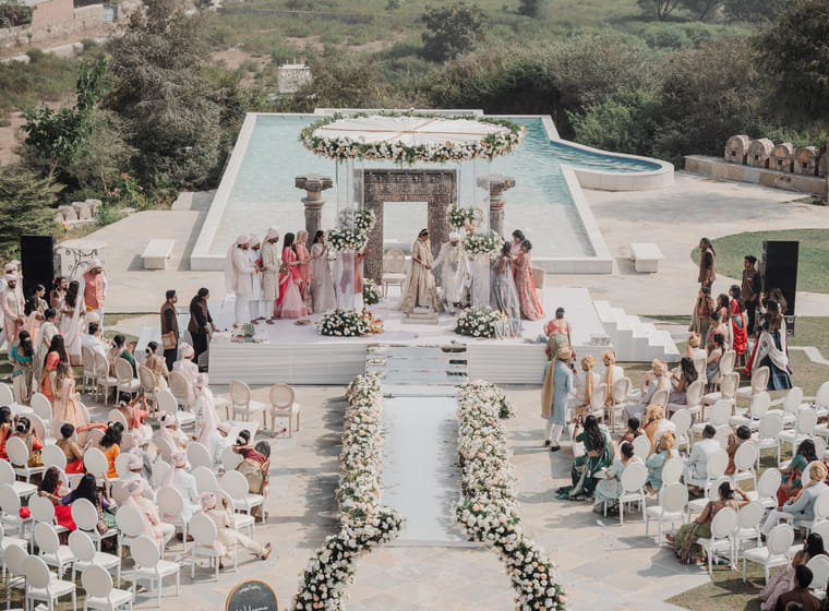 An elevated view of a poolside wedding ceremony with a floral mandap, a white petal aisle and rows of seated guests