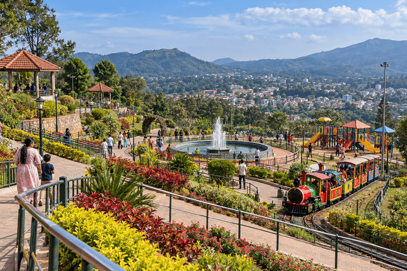 Flower-lined paths, a fountain, and a toy train at Mahatma Gandhi Park, with hills beyond