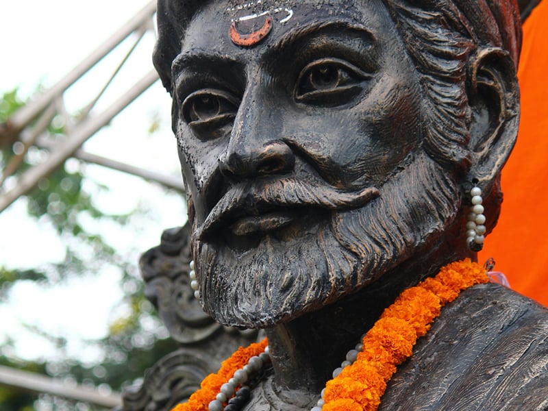 Bronze bust of Chhatrapati Shivaji Maharaj draped in a marigold garland, saffron banner behind | Shirgaonkar Beach Resort