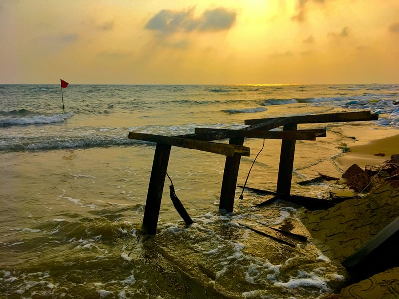 Weathered wooden frame standing in the shallows at sunset, with a red flag on the shore | Shirgaonkar Beach Resort