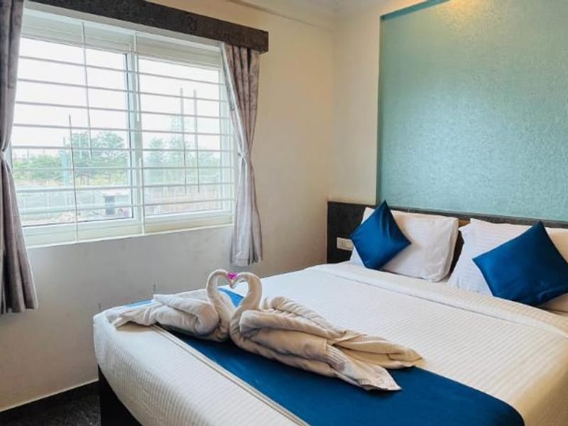 Queen bed with white linen, blue cushions, and a towel swan in the Standard AC Room at Value Inn, Bangalore Airport beside a bright window