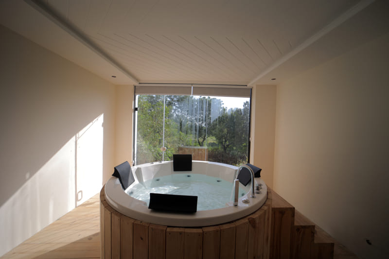 A wooden-framed Jacuzzi tub at Nazaara Hills Retreat, Udaipur, positioned beside a window with a forest view.