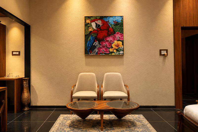 Two cream armchairs and a round teak table beneath a bright painting of macaws at The Garud Forest, Tadoba