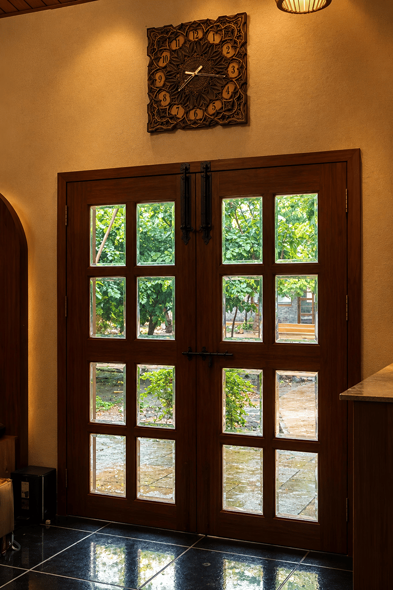 Wooden French doors opening onto greenery beside a carved wall clock in the lobby of The Garud Forest, Tadoba