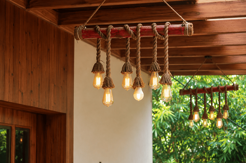 Edison bulbs hung on ropes from a bamboo pole beneath a timber ceiling, with green trees beyond at The Garud Forest, Tadoba