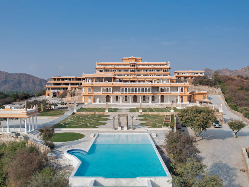 Palace-style hotel exterior with a central swimming pool, landscaped gardens, stone pathways, pavilion structures, and surrounding hills under a clear blue sky - Fateh Vilas, Udaipur