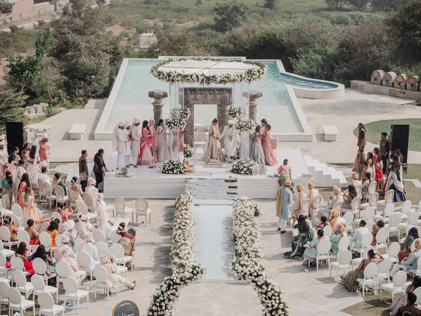 A wedding ceremony at the outdoor featuring as aisle decorated with flowers on either side, guests seated on either side and a centre stage where the rituals are taking place with bride, broom, and their close family members.