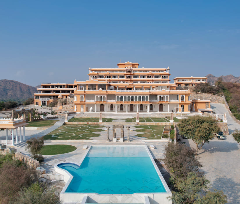 Palace-style hotel exterior with a central swimming pool, landscaped gardens, stone pathways, pavilion structures, and surrounding hills under a clear blue sky - Fateh Vilas, Udaipur