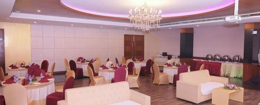 A banquet hall in Dhanalakshmi Srinivasan Hotels, Perambalur, with round tables, chairs, sofas, and a floral arrangement in the foreground. The hall has wooden flooring and a chandelier.