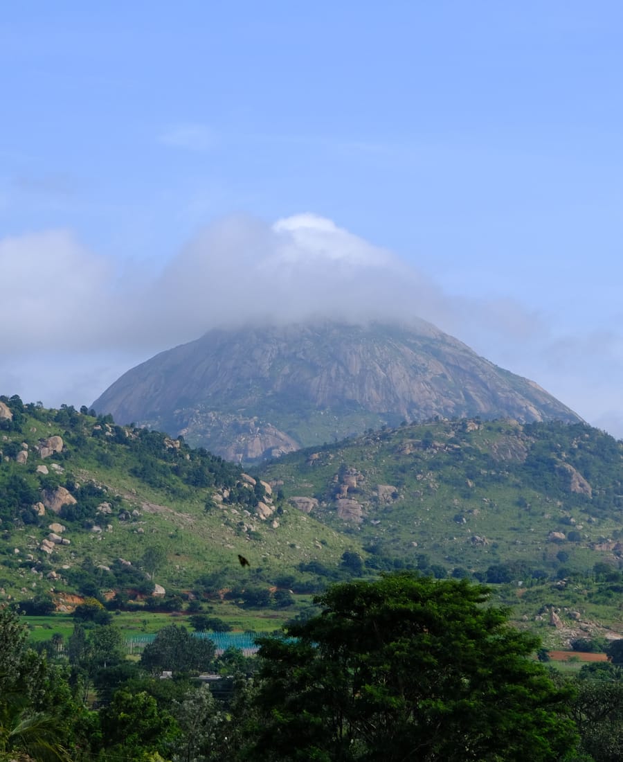 Nandi Hills near Bangalore, with the rocky granite peak capped by a cloud above green slopes and scattered boulders under a pale blue sky