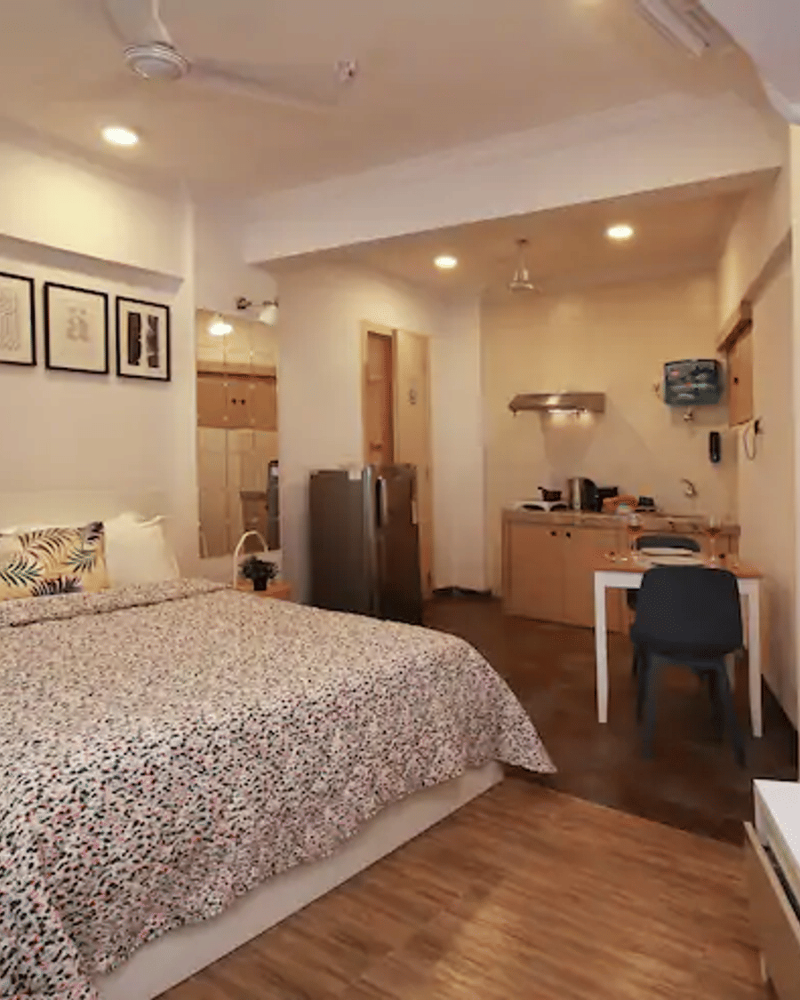 Bedroom of Room 301 at Wabi-Sabi, Juhu, with a printed duvet, wooden flooring, a wall-mounted TV showing the Netflix logo, and an open kitchenette beyond.
