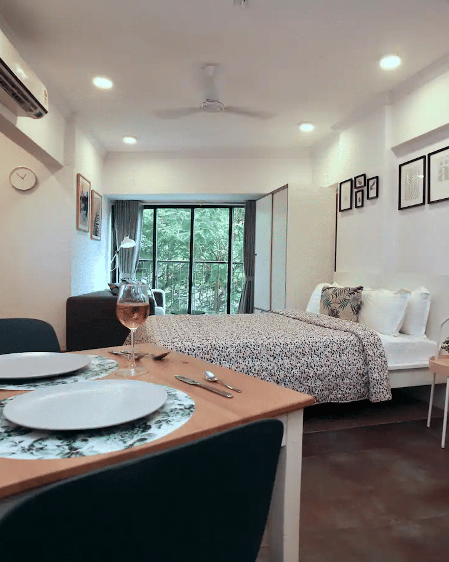 Bedroom of Studio Rooms (A) at Wabi-Sabi, Juhu, with a patterned bedspread, a mirrored wall, framed art, and a dining table with 2 place settings and wine glasses.