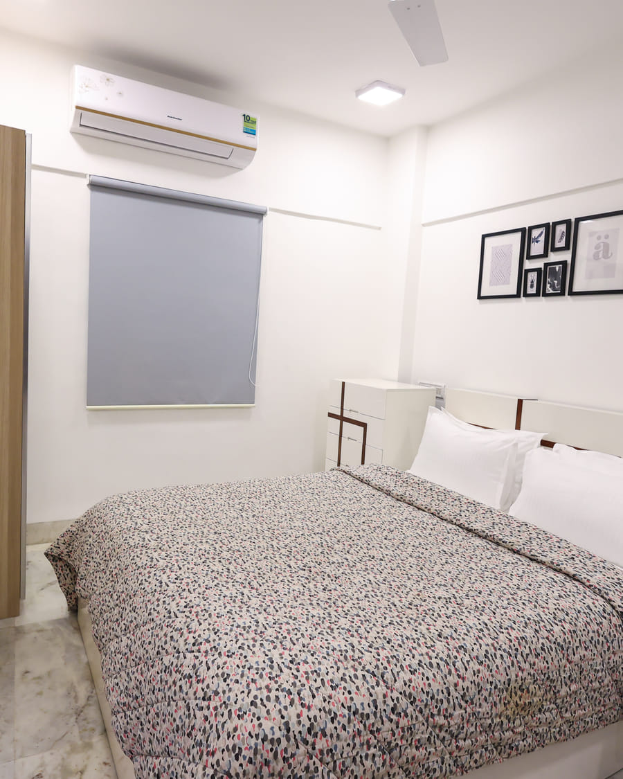 A wide view of the Room 401 bedroom at Wabi-Sabi, Juhu with a patterned bedspread, a split air-conditioning unit, a grey roller blind, and an open wooden wardrobe.