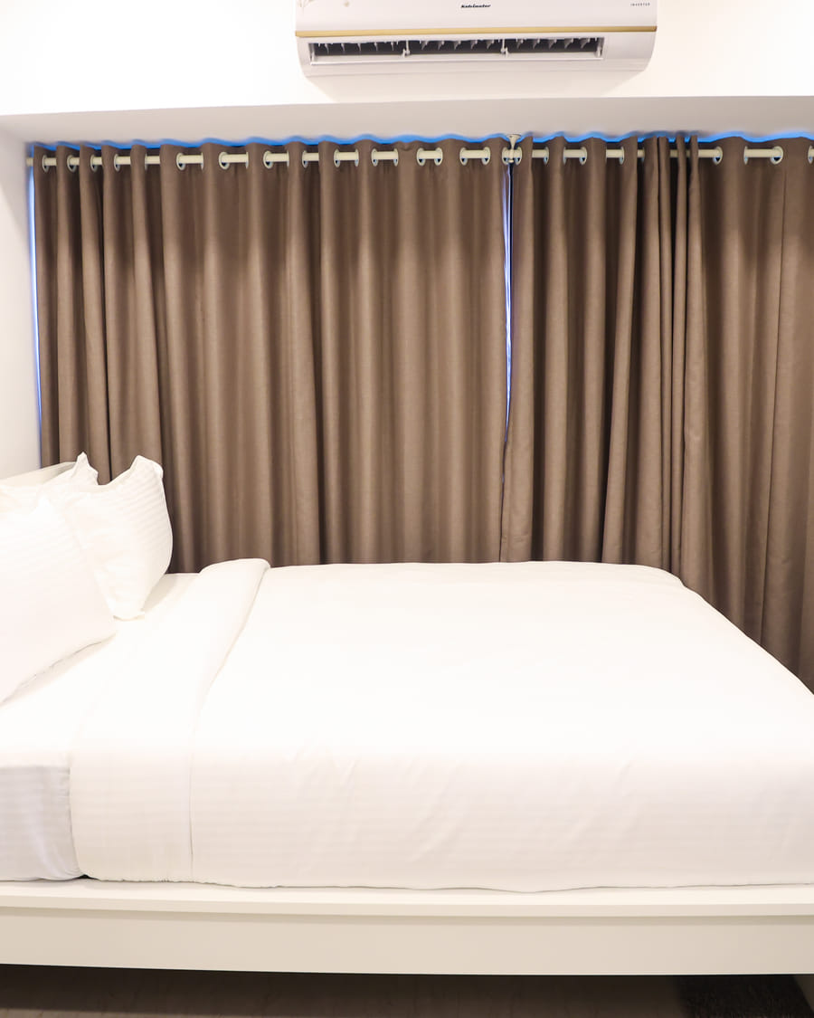 Bedroom of Studio Rooms (E) at Wabi-Sabi, Juhu, with a white bed, brown curtains, a wall-mounted AC, a gallery wall of framed prints, and a teal accent chair.
