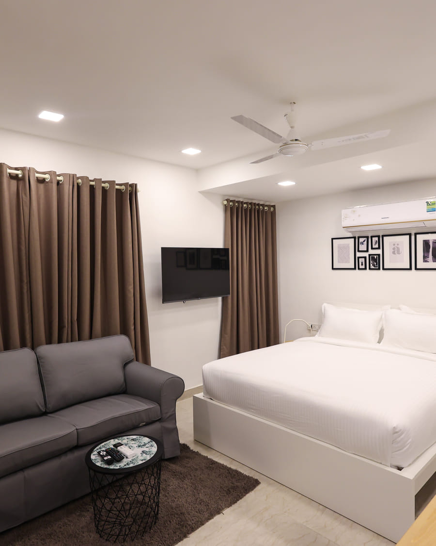 The Room 404 studio at Wabi-Sabi, Juhu with a white bed, wall-mounted television, grey sofa, round coffee table, and mirrored wardrobe.