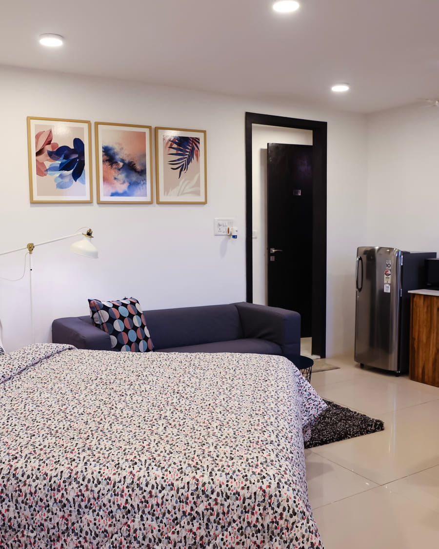 Studio Rooms (B) at Wabi-Sabi, Juhu, with a patterned bedspread, a navy sofa, framed art, and a kitchenette with a fridge, microwave, and dining table.