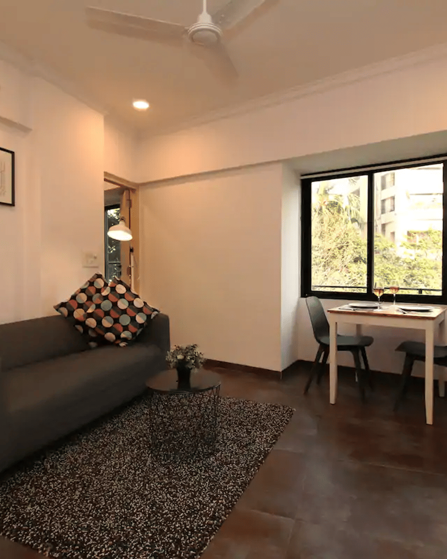 Living room of the 1BHK apartment at Wabi-Sabi, Juhu, with a grey sofa, framed art, a TV console, and a 2-seater dining table beside a window with wine glasses.