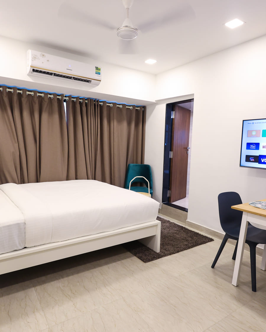 Bedroom at Wabi-Sabi, Juhu, with a bed, brown curtains, a wall-mounted AC, a smart TV showing streaming apps, and a dining table with 2 chairs and wine glasses.