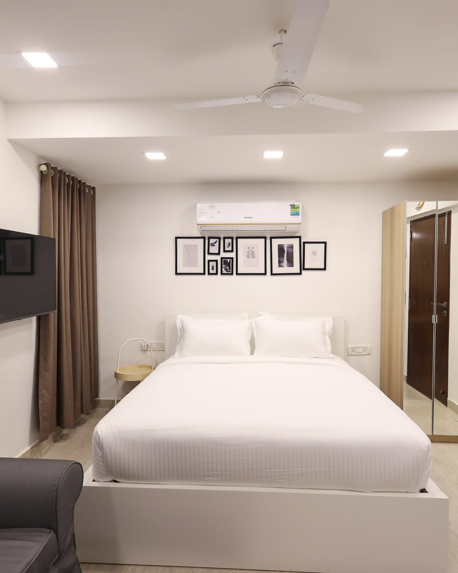A white bed beside a mirrored wardrobe in a Wabi-Sabi, Juhu bedroom, with framed wall art, a wall-mounted TV, a grey sofa and an AC unit.