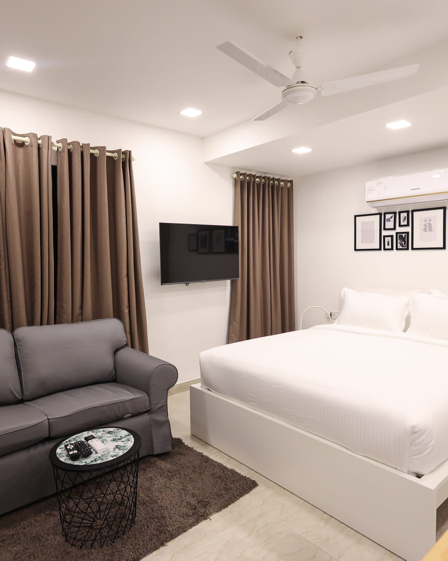 Bedroom at Wabi-Sabi, Juhu, with a white bed, a wall-mounted AC, a gallery wall of framed prints, a wall-mounted TV, and a 2-seater grey sofa.