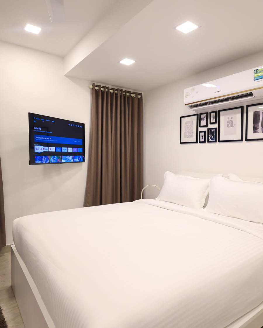 A grey armchair by brown curtains in a Wabi-Sabi, Juhu bedroom, with framed art on the gallery wall and a smart TV on its setup screen.