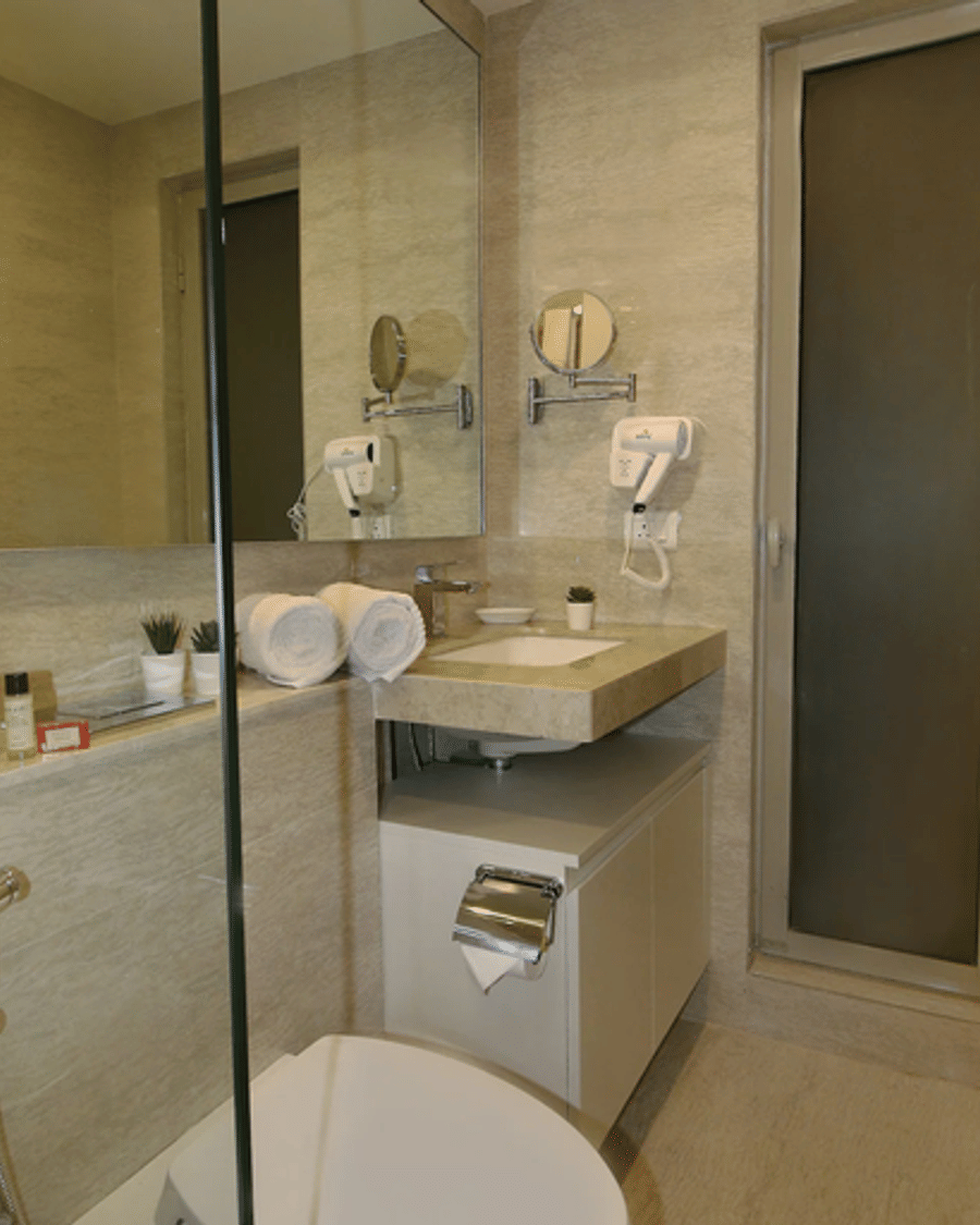 A Wabi-Sabi, Juhu bathroom with a vanity mirror, hair dryer, folded white towels, a wall-mounted shower, and a frosted-glass sliding door.