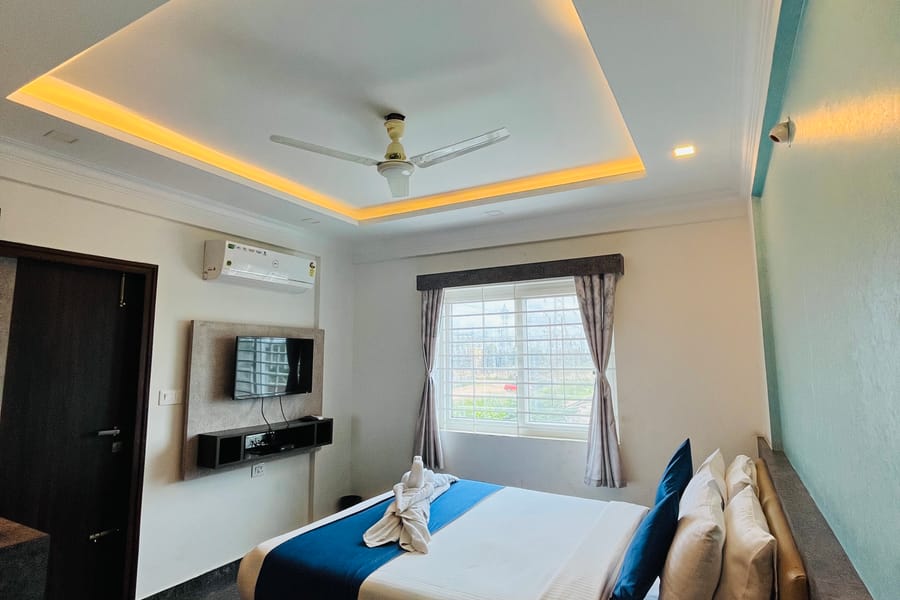 Queen bed under a recessed ceiling with warm cove lighting and a wall mounted TV in the Standard Balcony Room at Value Inn, Bangalore Airport