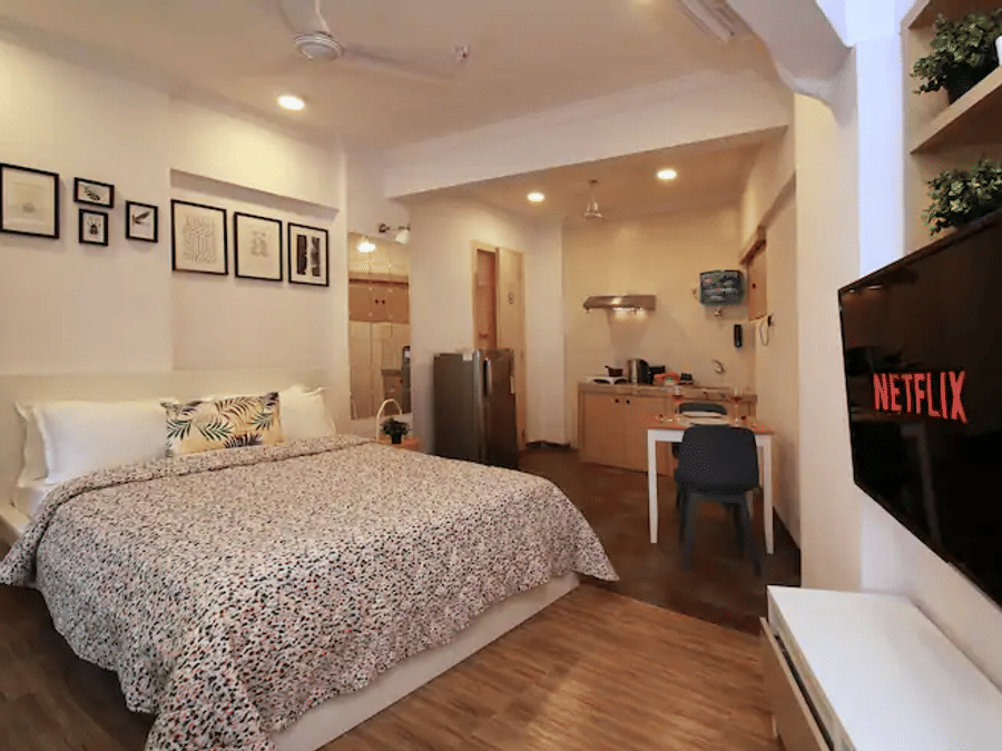 Bedroom of Room 301 at Wabi-Sabi, Juhu, with a printed duvet, wooden flooring, a wall-mounted TV showing the Netflix logo, and an open kitchenette beyond.