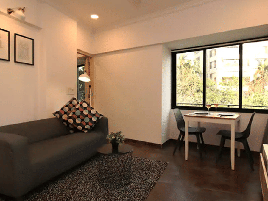 Living area at Wabi-Sabi, Juhu, with a grey sofa, a round coffee table, framed wall art, and a 2-seater dining table set beside a large window with a tree view.