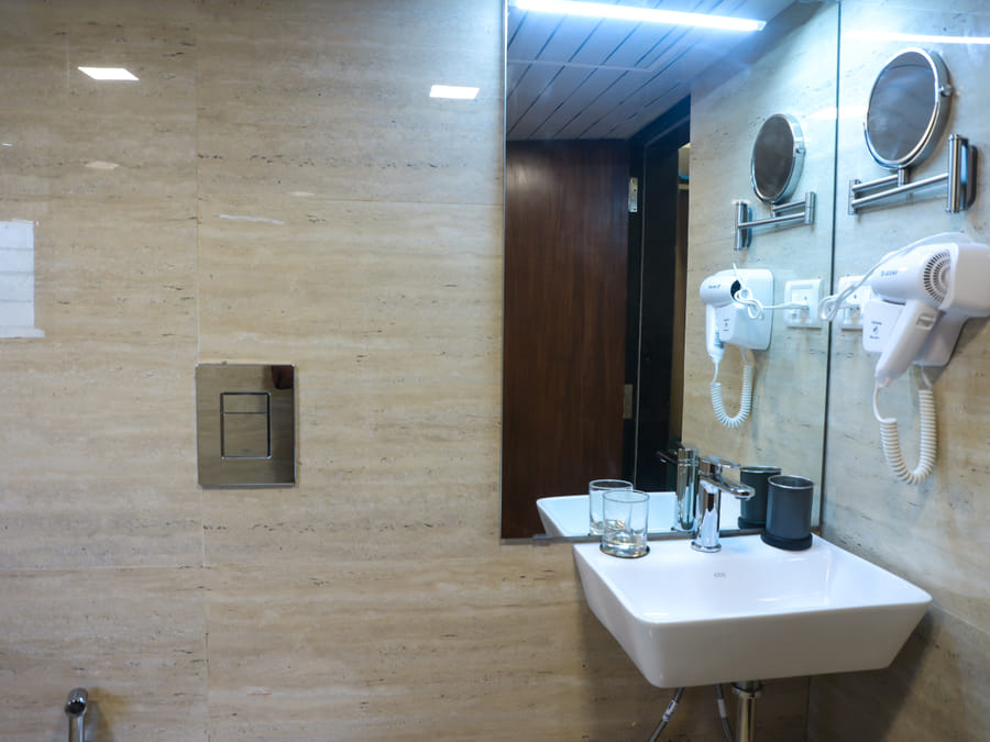 Bathroom of Rooms 402 and 5001 at Wabi-Sabi, Juhu, with a beige travertine tile wall, a white sink, a wall-mounted hairdryer, and a swivel mirror.
