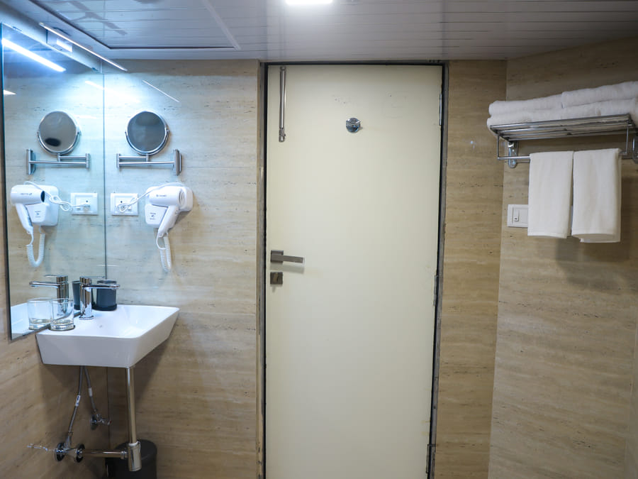 Bathroom of Rooms 402 and 5001 at Wabi-Sabi, Juhu, with the entry door, a towel rack of folded white towels, and the mirrored sink counter.