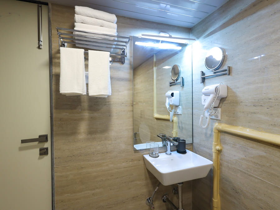 Bathroom of Rooms 404 and 5003 at Wabi-Sabi, Juhu, with a towel rack of folded towels, an exposed yellow pipe, and a mirrored sink with a hairdryer.