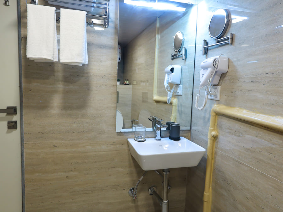 Bathroom of Rooms 404 and 5003 at Wabi-Sabi, Juhu, viewed straight-on at the sink, with the toilet reflected in the mirror and an exposed yellow pipe.