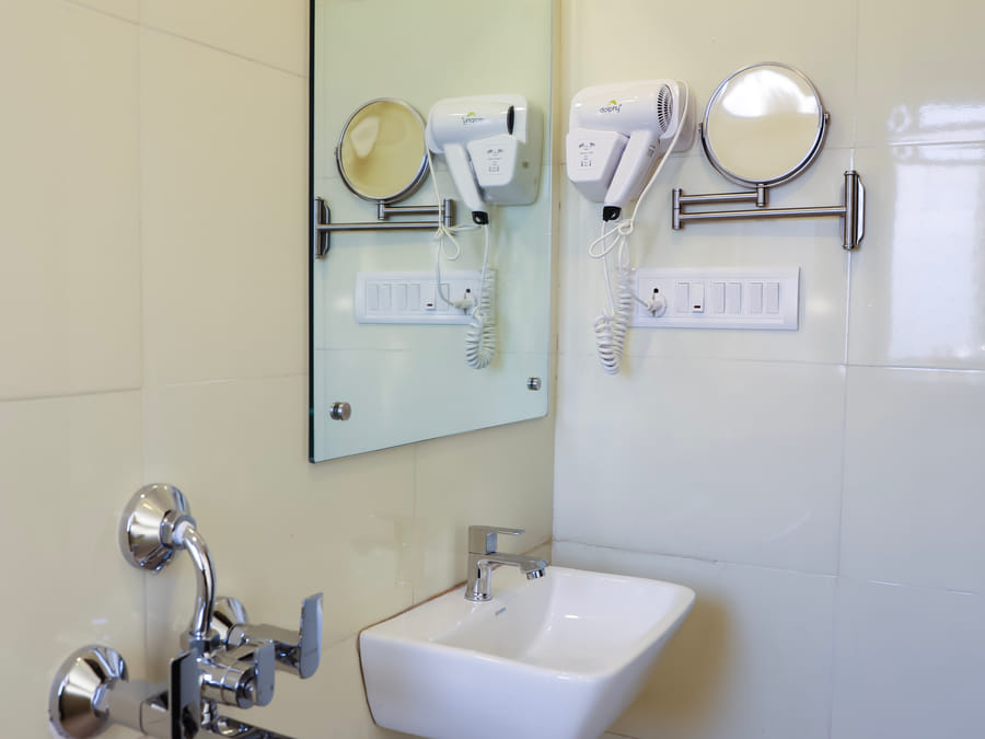 Bathroom of Room 501 at Wabi-Sabi, Juhu, with cream square wall tiles, a white sink, a wall-mounted hairdryer, a swivel mirror, and an exposed tap valve.