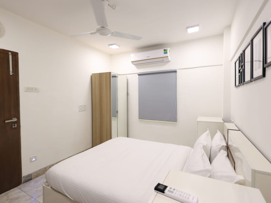 Bedroom of Room 401 at Wabi-Sabi, Juhu, showing a mirrored wardrobe, a grey roller blind, an air conditioner, a wall clock, and a wooden entry door.
