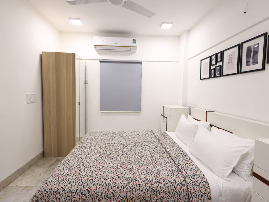 The Room 401 bedroom at Wabi-Sabi, Juhu with a patterned bedspread, white pillows, a wooden wardrobe, a grey roller blind, and a gallery of framed pictures on the wall.