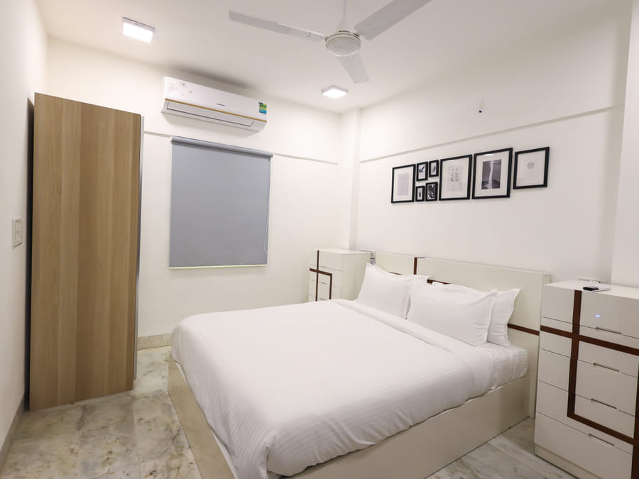 Bedroom of Room 401 at Wabi-Sabi, Juhu, with a closed wooden wardrobe door, a grey roller blind, and a full view of the bed on its raised platform.