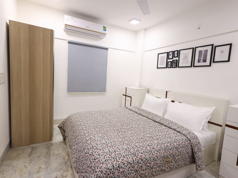 A wide view of the Room 401 bedroom at Wabi-Sabi, Juhu with a patterned bedspread, a split air-conditioning unit, a grey roller blind, and an open wooden wardrobe.
