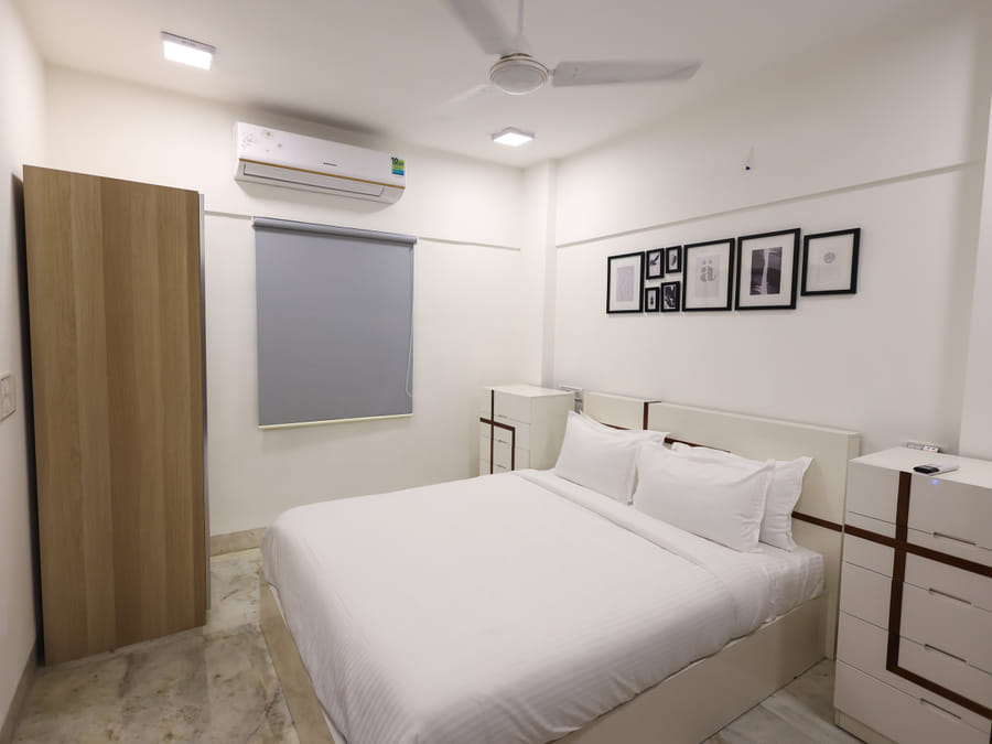 Bedroom of Room 401 at Wabi-Sabi, Juhu, with an open plain wooden wardrobe door, a single grey roller blind, and a gallery wall of framed art.