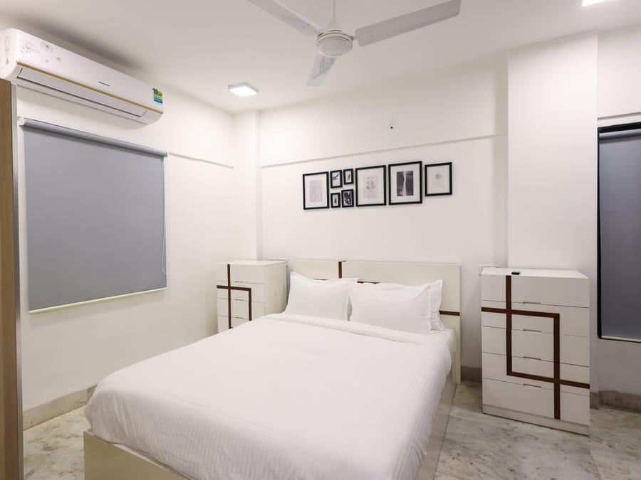 Bedroom of Room 401 at Wabi-Sabi, Juhu, with a grey roller blind, a gallery wall of framed art, and two dressers with a brown geometric inlay.