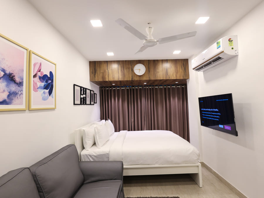 Bedroom of Rooms 402, 3001, and 5001 at Wabi-Sabi, Juhu, with a wall-mounted TV showing a Netflix screen, a grey sofa, and a wooden panel clock.