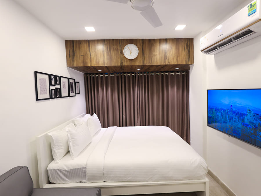 Bedroom of Rooms 402, 3001, and 5001 at Wabi-Sabi, Juhu, with a wall-mounted TV showing a city skyline screensaver and a wooden panel clock above the bed.